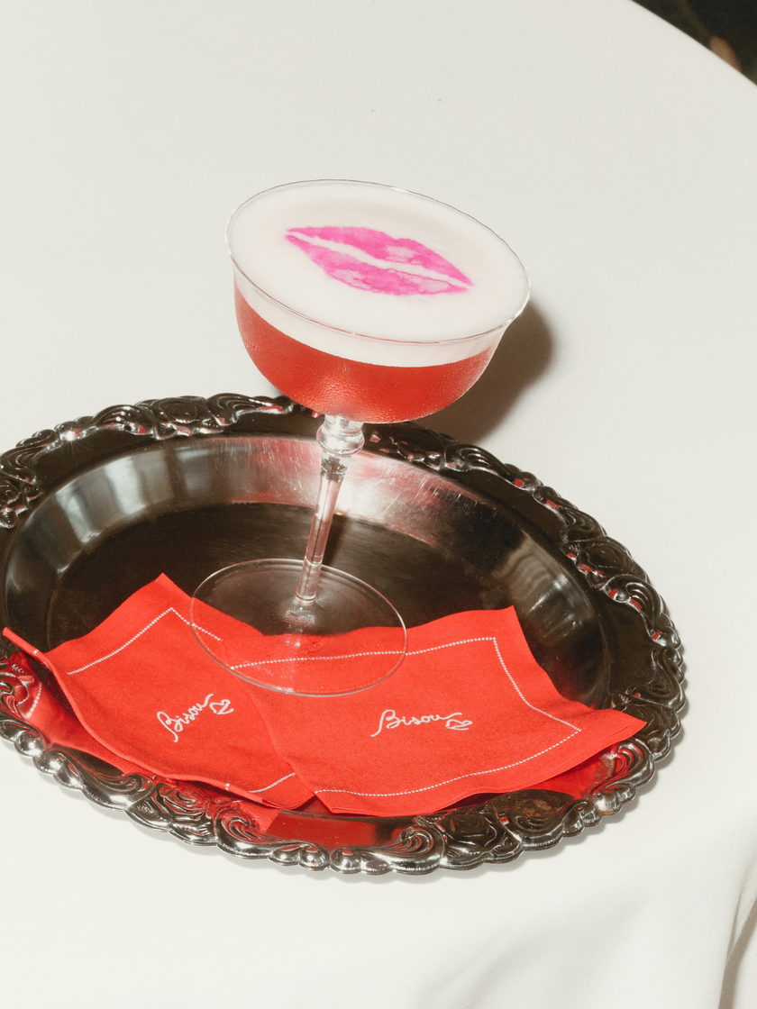 A Bisou Bisou cocktail on a silver tray — a coupe of red drink under a white foam, printed with a pink lipstick kiss, on Bisou's red monogrammed napkins.